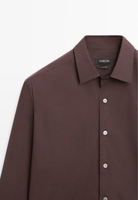 Chemise marron foncé à manches longues avec boutons, col pointu, présentant quatre boutons blancs visibles et une étiquette de marque à l'intérieur du col.