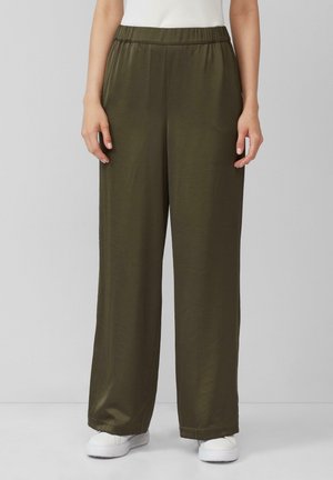 Femme portant un pantalon large vert olive avec une taille élastique et des baskets blanches, debout devant un fond uni gris clair.