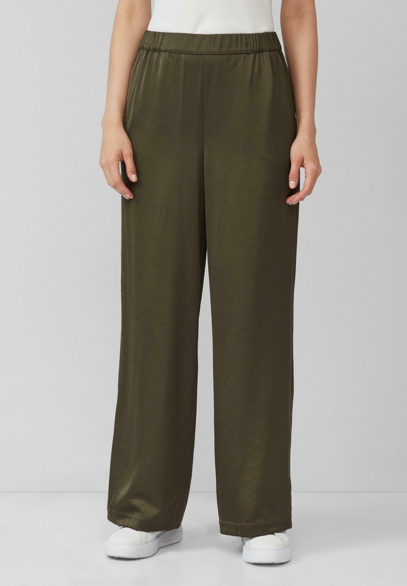 Femme portant un pantalon large vert olive avec une taille élastique et des baskets blanches, debout devant un fond uni gris clair.