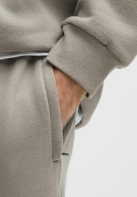 Hand van een persoon in een beige gebreide broekzak, met een bijpassende beige gebreide trui met geribbelde manchetten, tegen een effen lichte achtergrond.