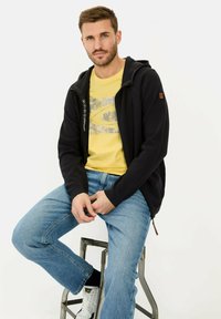 Sort zip-up hættetrøje lavet af tekstureret stof, med hætte, sidelommer og logo detaljer, parret med en gul t-shirt og blå jeans.