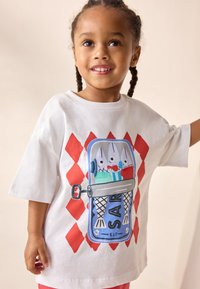 Fehér pamut póló, nagy grafikával, amely egy konzervdobozban lévő rajzfilmhalakat ábrázol kék és piros részletekkel, rövid ujjú és laza szabású.