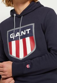 Sudadera con capucha azul marino con un gran logo en forma de escudo que presenta rayas verticales rojas y blancas, contorno gris y texto "GANT" en negrita blanca. Incluye cordones ajustables.