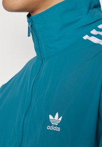 Žalias zip-up džemperis su aukšta apykakle ir trimis baltomis juostelėmis dešinėje peties pusėje. Adidas logotipas siuvinėtas ant kairiojo krūtinės.