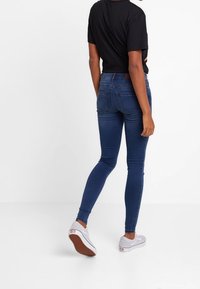 Jeans ajustados de denim azul oscuro, con cintura de altura media, diseño estándar de cinco bolsillos, combinados con zapatillas blancas y una camiseta negra.