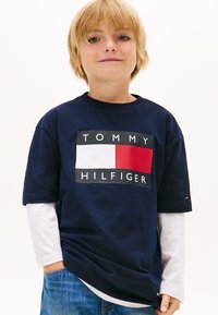 Tricou din bumbac bleumarin, cu un grafic mare format din dreptunghiuri roșii și albe și textul „TOMMY HILFIGER”. Purtat peste un tricou cu mâneci lungi alb.