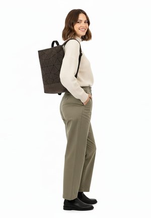 Mujer de pie sonriendo, con suéter beige, pantalones oliva, botas negras y una mochila negra con patrón geométrico sobre un hombro.