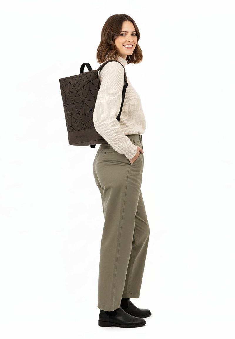 Vrouw staat en glimlacht, draagt een beige trui, olijfgroene broek, zwarte laarzen en een zwarte rugzak met geometrisch patroon over één schouder.