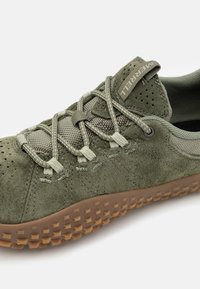 Zapato verde de ante con secciones de malla, superficie texturizada y suela de goma con pequeños patrones cuadrados elevados. Los cordones están pasados a través de ojales.