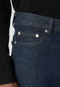Mørkeblå denimjeans med rett ben, med sølvriveter, kontrastsøm og middels høy midje. Teksturen er glatt og robust.