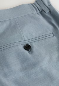 Détail d'un pantalon de costume bleu clair avec un unique bouton noir et une poche arrière horizontale soulignée de coutures décoratives.