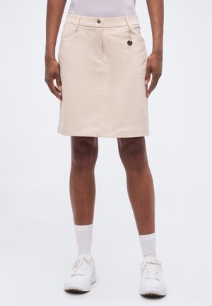 Osoba nosiaca béžovú sukňu po kolená s prednými vreckami, kovovým gombíkom a malým okrúhlym príveskom, v kombinácii s bielymi ponožkami a topánkami.