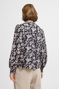 Blusa a maniche lunghe nera con motivo floreale bianco, con bottoni sul retro, con maniche arricciate e un fit morbido, abbinata a pantaloni beige.