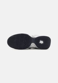 Semelle de chaussure de sport blanche et argentée avec une semelle extérieure en caoutchouc noir présentant un motif en chevrons et un logo en relief. Texture lisse, forme arrondie.