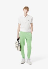 Hvid polo-shirt med et lille grønt logo, parret med lysegrønne sweatpants og hvide sneakers, holder en beige taske med sorte detaljer.