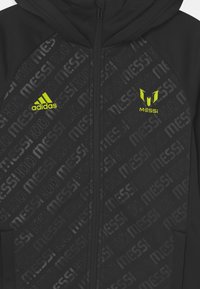 Giacca nera con cappuccio e chiusura con zip, caratterizzata da un motivo geometrico e branding "MESSI" in una finitura lucida e tonale. Dettagli in giallo brillante.