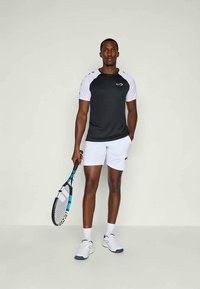 Camiseta atlética em preto e branco com mangas curtas e logótipo, combinada com calções brancos e ténis, segurando uma raquete de ténis.