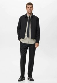 Črna jakna z ovratnikom, siva srajca z gumbi, črni sukanec in črne hlače. Model nosi naoljene črne čevlje.