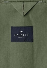 Zelena tkanina s pravokutnom tamnoplavošom etiketa prišivenom na nju, prikazujući natpis "Hackett London" u bijelom tekstu i logotipom prekriženih vesala iznad.