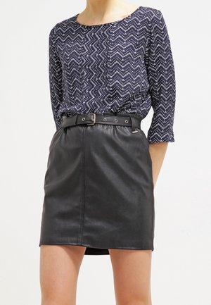 Femme portant un haut bleu marine à motif en zigzag avec des manches trois-quarts, rentré dans une mini-jupe en cuir noire ceinturée.