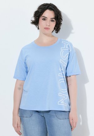 Femme aux cheveux courts bouclés et foncés porte un T-shirt bleu clair avec un texte blanc et un jean bleu, debout contre un mur blanc avec une ombre.