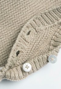 Dettaglio di un maglione lavorato a maglia beige con orlo a coste, chiusure con bottoni e motivi ad occhio, realizzato in morbida lana o filato acrilico.