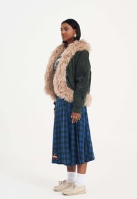 Giacca bomber verde con colletto in pelliccia eco-beige, abbinata a una gonna midi a scacchi blu e scarpe beige, con un accento decorativo.