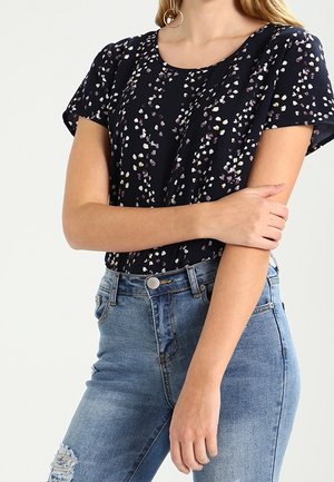 Femme portant un haut noir à manches courtes avec de petits motifs multicolores et un jean bleu clair déchiré, tenant un bras croisé sur son corps.