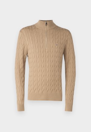Beige Kabelstrickpullover mit hohem Kragen und halbem Reißverschluss. Mit gerippten Bündchen und Saum, zeigt ein kleines Logo auf der Brust.