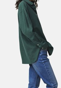 Waldgrünes Langarmshirt mit Kragen, zwei Knöpfen am Ärmelsaum und legerem Schnitt, kombiniert mit blauen Jeans.