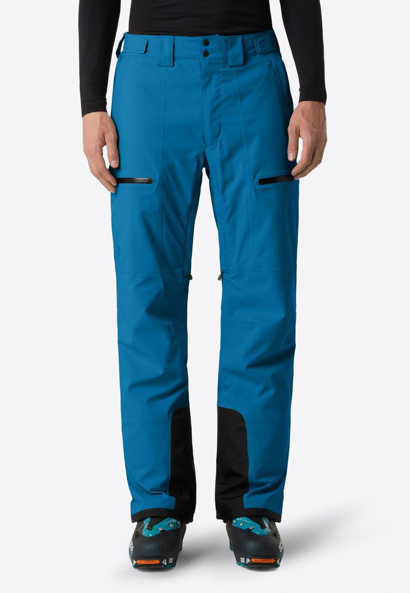 Pantaloni da esterno blu con polsini rinforzati neri, tasche sulla coscia con zip, indossati con scarponi da sci e una maglia nera a maniche lunghe.