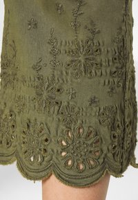 Olive green fabric skirt with floral embroidery and cut-out detailing at the hem, featuring a scalloped edge and textured finish.