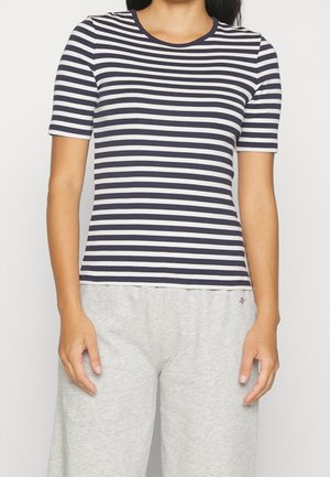 Femme portant un t-shirt à manches courtes ajusté à rayures horizontales bleu marine et blanches, et un pantalon ample gris clair, debout devant un fond uni.
