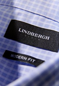 Zawieszki na kołnierzu jasnoniebieskiej koszuli w kratę. Zawieszki mają napisy „LINDBERGH BLACK” oraz „MODERN FIT” na czarnym materiale z białym tekstem.