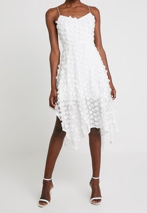 Kobieta ubrana w białą, bezrękawną sukienkę z aplikacjami 3D w kształcie kwiatów i asymetrycznym plisowanym dołem, zestawioną z białymi szpilkami z paskiem wokół kostki, na jednolitym tle.