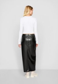Zwarte leren midi-jurk met achterzakken en subtiele stikdetails, gecombineerd met een witte top met lange mouwen en beige sneakers.