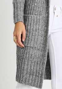 Szary sweter rozpinany na drutach o fakturze prążkowanej, z przednią kieszenią i długimi rękawami, noszony na białych spodniach. Widoczny jest miękki, gruby splot włóczki.