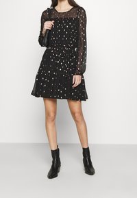 Vestido negro con mangas largas de tul, con un diseño en capas, acentos metálicos dispersos y una cintura ajustada con un detalle de lazo.