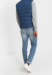 Marinblå puffy väst över en grå långärmad skjorta, ihop med ljusblå jeans och vita sneakers, sedd bakifrån.