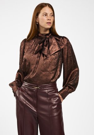 Vrouw in een glanzende bronzen blouse met lange mouwen en een grote strik, gecombineerd met hoogdoorgetrokken, donkerbruine leren broek. Ze heeft haar handen in haar zakken en kijkt naar rechts.