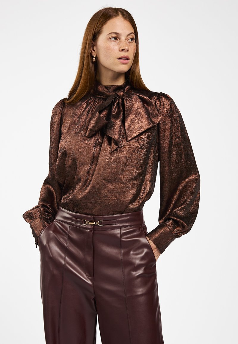 Vrouw in een glanzende bronzen blouse met lange mouwen en een grote strik, gecombineerd met hoogdoorgetrokken, donkerbruine leren broek. Ze heeft haar handen in haar zakken en kijkt naar rechts.