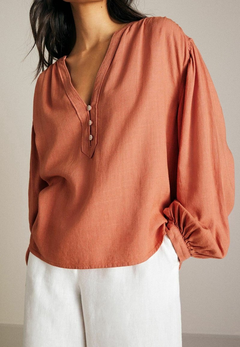 Femme portant un chemisier ample couleur rouille avec trois boutons et un pantalon blanc, visible des épaules aux hanches sur un fond uni.