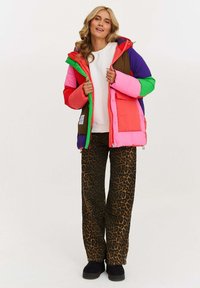 Farebná nafukovacia bunda s panelmi v ružovej, zelenej, purpurovej a hnedej farbe. Nosí sa cez bielu košeľu s nohavicami v leopardom vzore a čiernymi topánkami.