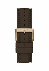 Brūns silikona pulksteņa siksniņa ar teksturētām malām, misiņa krāsas metāla sprādze ar gravējumu "Gc" un "Gc" logotips uz siksniņas cilpas.