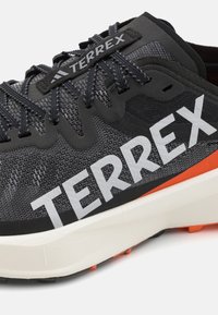 Svart och grå löparsko med en strukturerad mesh-överdela, fet vit "TERREX"-logotyp, orange accent och en vadderad vit mellansula.