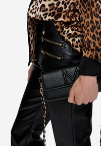 Borsa a tracolla in pelle nera con superficie texturizzata, caratterizzata da una tracolla in catena dorata, appoggiata su una maglietta con stampa leopardata e pantaloni in pelle nera.