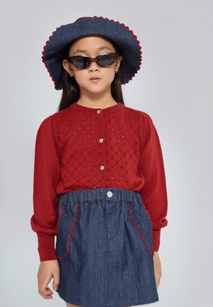 Cárdigan rojo de punto con un patrón calado y botones al frente. Falda vaquera con costuras rojas y bolsillos. Sombrero azul con borde festoneado. Gafas de sol negras.