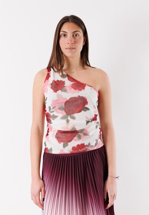 Donna con capelli castani lisci indossa un top bianco a una spalla con stampa floreale rossa e una gonna plissettata in ombré dal viola al rosa, in piedi davanti a uno sfondo bianco.