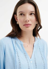 Blusa azzurro chiaro realizzata in tessuto testurizzato con pois, con un inserto in pizzo lungo la patta dei bottoni e un colletto parziale.