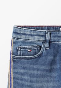 Pantalones vaqueros de mezclilla con un lavado claro, que presentan un pequeño bolsillo frontal y rayas laterales en azul y dorado metálico, acentuados por un logo en rojo, blanco y azul.